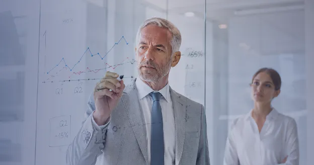 A senior executive writing performance charts on a glass board, a colleague in the background