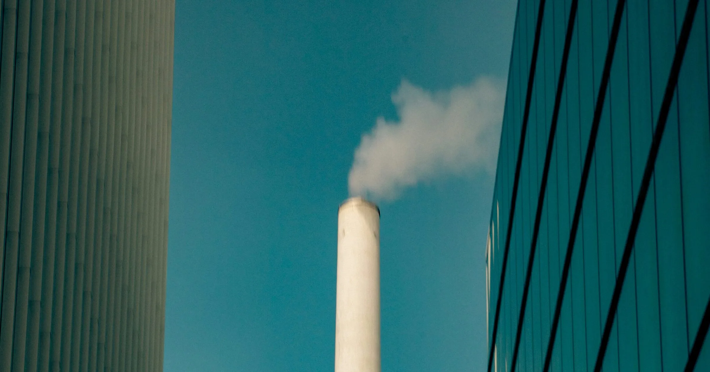 An industrial smokestack rising between a concrete office block and a glass curtain wall